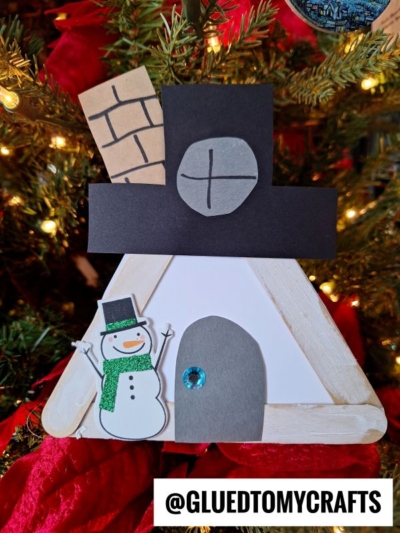 A handmade popsicle stick snowman house, crafted from sticks and paper, features a snowman, a door with a blue gem, and a chimney. Set before a glowing Christmas tree, it brings festive charm to your holiday décor.