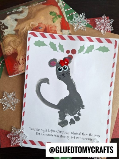 A Christmas-themed craft features a charming christmas mouse made from a handprint, decorated with googly eyes, a red pompom nose, and holly leaves on top, sitting on striped border paper with a book and snowflake ornaments nearby.