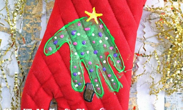 A red potholder features a painted handprint Christmas tree, decorated with colorful dots as ornaments and topped with a yellow star. Gold tinsel is scattered in the background.
