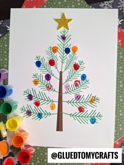 A thumbprint Christmas tree made from green painted lines for branches, brown paper for the trunk, and colorful fingerprint ornaments on white paper. A gold star tops the tree, with paint pots and patterned paper surrounding the festive artwork.