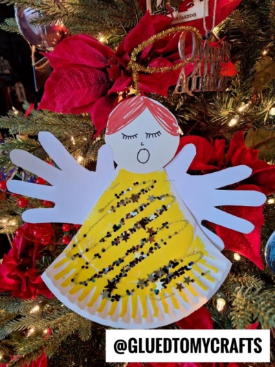 A handmade paper plate handprint angel ornament with yellow and black glitter on its dress, handprint wings, and red hair hangs on a Christmas tree decorated with red poinsettias. @GLUEDTOMYCRAFTS is written at the bottom.