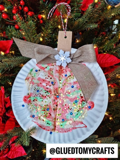 A colorful, glittery holiday craft inspired by paint splat christmas ornaments hangs on a tree. Made from a coffee filter and paper plate, it's decorated with sequins, a burlap bow, a felt snowflake, and gift tag among red flowers and pine needles.
