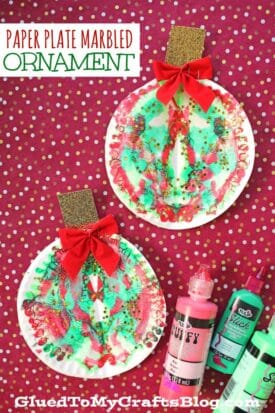 Two colorful marble Christmas ornaments made from paper plates, decorated with red bows and gold tops, are displayed on a red polka dot background. Craft paint bottles are nearby. Text reads, Paper plate marbled ornament.