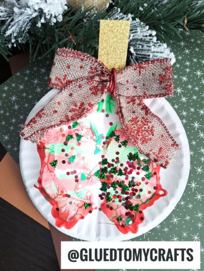 A paper plate paint splat Christmas ornament craft decorated with red and green paint blots, glitter, a large gold and red bow, and a gold ornament top. The background features a green patterned surface and pine branches.