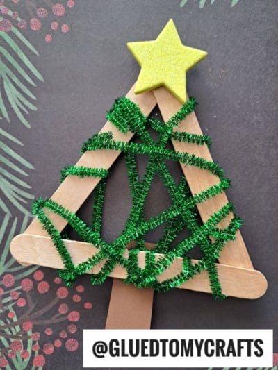 A pipe cleaner Christmas tree craft made from popsicle sticks shaped into a triangle, decorated with a yellow star on top and green pipe cleaners wrapped around it. @GLUEDTOMYCRAFTS is written at the bottom.