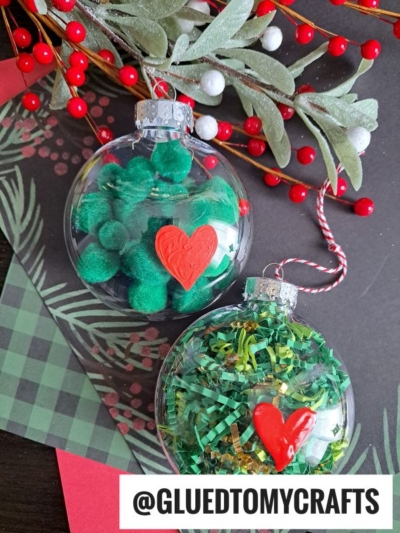 Two clear Christmas ornaments filled with green pom-poms and tinsel, each decorated with a red heart for a charming Grinch pom pom ornament look. Festive greenery and red berries surround the ornaments. Text at the bottom reads @GLUEDTOMYCRAFTS.