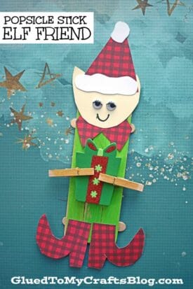 A popsicle stick elf craft featuring a red and green outfit, holding a small present. The smiling elf has googly eyes, a red plaid hat and boots, all set against a blue background with gold star decorations.