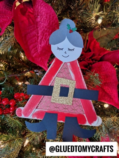 A handmade Popsicle Stick Mrs. Claus paper craft ornament with a red dress and gold belt hangs on a Christmas tree decorated with red poinsettias and berries. @GLUEDTOMYCRAFTS is written at the bottom.