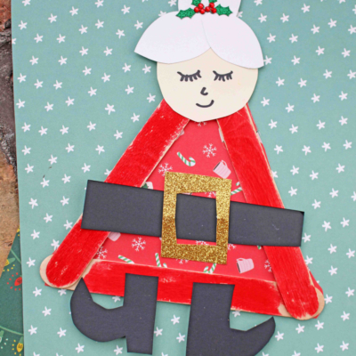 Create a festive Popsicle Stick Mrs. Claus with red sticks, a paper face, white hair bun with holly, glittery gold belt, black boots, and a red patterned dress—all set against a blue snowflake background.