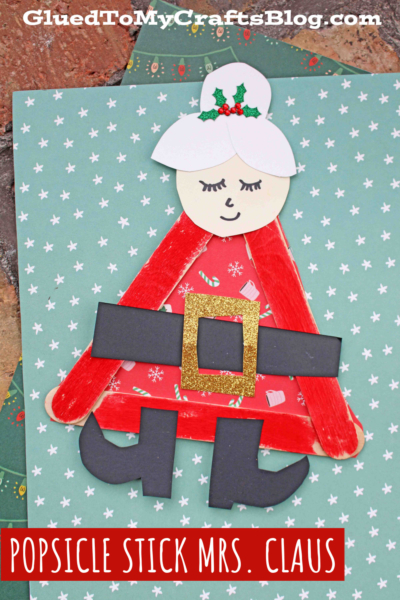 Create a festive Popsicle Stick Mrs. Claus with red sticks, a paper face, white hair bun with holly, glittery gold belt, black boots, and a red patterned dress—all set against a blue snowflake background.