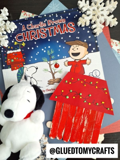 A Charlie Brown Christmas book, a Snoopy plush, snowflake decorations, and layered colored paper alongside a popsicle stick Snoopy's dog house decorated with Christmas lights. @GLUEDTOMYCRAFTS text at the bottom.