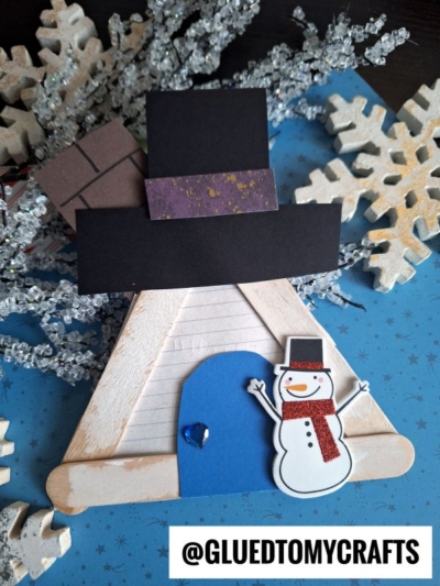 A handmade winter craft featuring a popsicle stick snowman house teepee, decorated with a paper snowman, a blue door with a blue heart gem, black chimney pieces, and snowflake ornaments on a blue background. Text: @GLUEDTOMYCRAFTS.