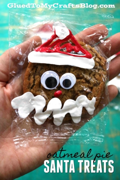 A hand holds an oatmeal cream pie gingerbread man, decorated with white and red icing to look like Santa’s face, with googly eyes and a red nose, wrapped in clear plastic. Text reads Gingerbread Man Treats SANTA TREATS.