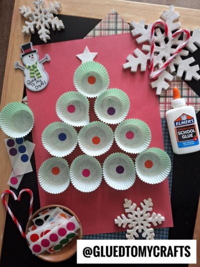 A cupcake liner Christmas tree craft made with green cupcake liners, colorful dot stickers, and a white star on red paper, surrounded by glue, candy canes, wooden snowflakes, and a snowman decoration.