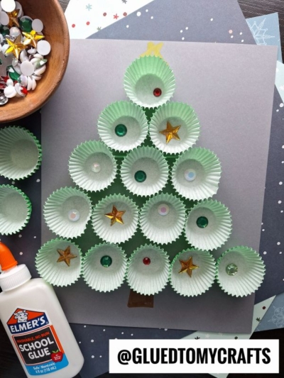 A cupcake liner Christmas tree, adorned with sequins and stars, is glued to gray paper. Elmer’s glue, a bowl of craft supplies, and papers are nearby. Text reads “@GLUEDTOMYCRAFTS.”.