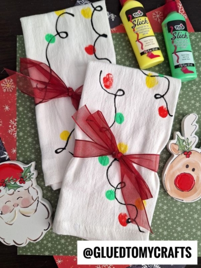 Two white towels decorated with colorful fingerprint Christmas lights napkins are tied with red ribbon bows. They rest on festive paper with Santa and reindeer cutouts, alongside red and yellow puffy paint bottles.