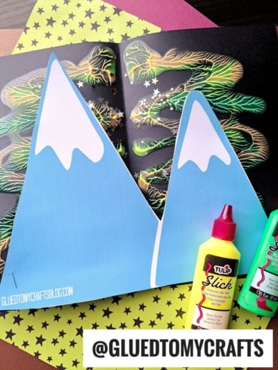Two blue paper mountains with snowy peaks sit on a craft background, surrounded by colorful star paper, neon paint bottles, and a marbled art piece featuring a paint splat northern lights effect. The handle @GLUEDTOMYCRAFTS is at the bottom.