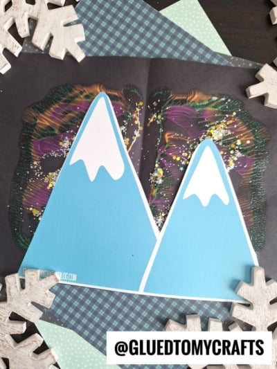 Two blue paper mountains with white snowy tops are placed over a vibrant paint splat evoking northern lights on black paper, resembling wings or a butterfly. Wooden snowflake decorations and patterned paper frame the colorful artwork.