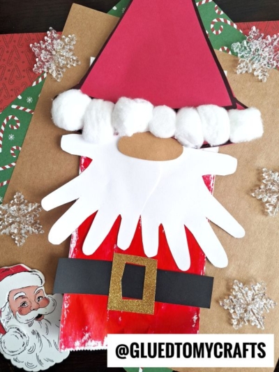 A Paper Bag Santa Puppet craft made with red, white, and black paper, cotton balls, a golden belt buckle, and cut-out handprints for the beard, placed on festive paper with snowflake decorations.