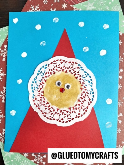 A handmade holiday card features a blue background with white dots, a red triangle, and a circle doily Santa face with googly eyes and a red nose. @GLUEDTOMYCRAFTS is at the bottom.