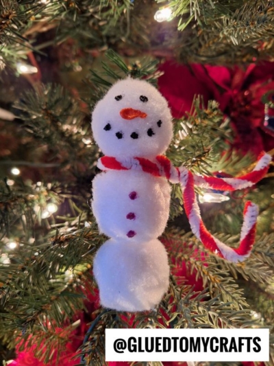 A handmade pom pom snowmen ornament with a red and white scarf hangs on a Christmas tree, decorated with lights. The snowman features white pom-poms, black eyes, mouth, buttons, and an orange nose. @GLUEDTOMYCRAFTS is written below.