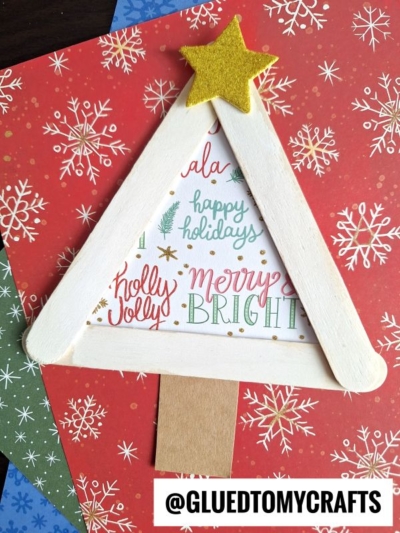 A handmade Christmas tree card features popsicle stick winter trees forming a festive shape, topped with a yellow felt star. Holiday text decorates the inside, and the card rests on red and green snowflake-patterned paper backgrounds.