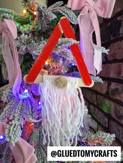 A Christmas tree adorned with a yarn bearded Santa ornament, crafted from red and beige craft sticks, white yarn for the beard, and a pompom nose. The festive handmade decoration hangs among snow-dusted branches and pink bows.