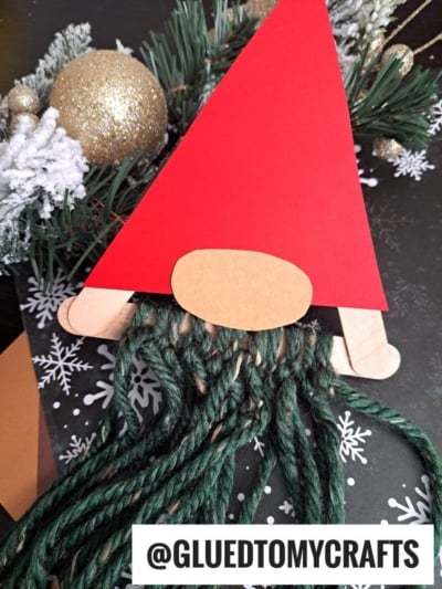 A yarn bearded Santa Christmas gnome craft made with a red paper hat, beige oval nose, popsicle stick feet, and green yarn beard, lying on a snowflake-patterned background with pine and gold ornaments. @GLUEDTOMYCRAFTS is visible.