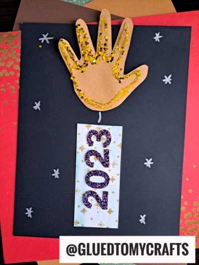 A handmade New Year’s craft featuring a brown handprint firework decorated with gold glitter, attached to a white rectangle with 2023 in glitter, on a black paper background with small star shapes.