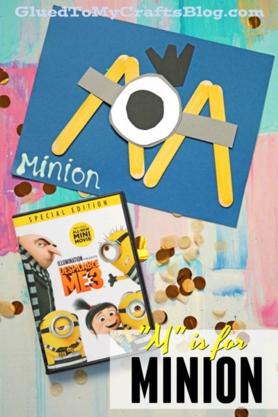 Craft project featuring a popsicle stick minion made from craft sticks and paper, displayed above a Despicable Me 3 DVD. Text reads M is for Minion. Confetti scattered on a colorful surface.