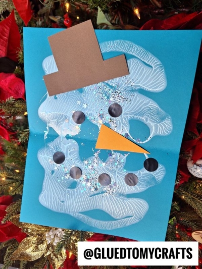 A paint splat melted snowman craft made from white paint on blue paper, decorated with black circle eyes and buttons, an orange triangle nose, a brown paper hat, glitter, and @GLUEDTOMYCRAFTS text at the bottom.