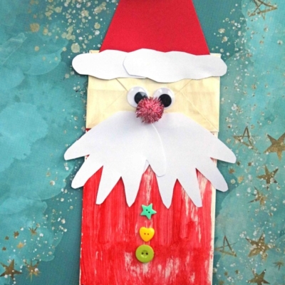 A handmade Paper Bag Santa Puppet features a red hat, white beard, googly eyes, pink pompom nose, and colorful buttons—crafted from a paper bag and set against a blue background with gold star designs.