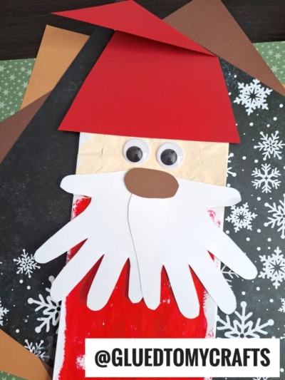 A Paper Bag Santa Puppet craft made from paper, featuring googly eyes, a white paper handprint beard, red hat, brown nose, and a snowy background. Text reads @GLUEDTOMYCRAFTS. Try this fun paper bag santa idea with your kids!.