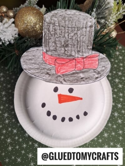 A paper plate snowman with a drawn-on black hat and red bow, orange carrot nose, and black dot smile. Festive greenery and gold ornaments are in the background—perfect inspiration for your next paper bowl snowman craft! Text reads @GLUEDTOMYCRAFTS.