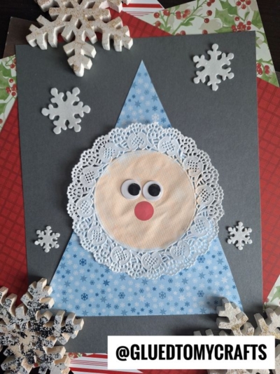 A handmade Christmas craft features a charming paper doily santa face with googly eyes and a red nose, topped with a blue triangle hat and surrounded by decorative snowflakes. @GLUEDTOMYCRAFTS is written at the bottom.