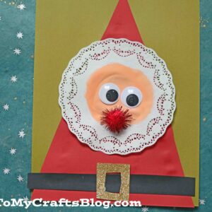 A handmade circle doily Santa craft featuring a paper doily, red triangle hat, googly eyes, red pom-pom nose, black belt with a glittery buckle—all set on a yellow background.