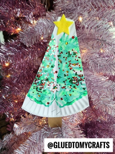 A paint splat Christmas tree made from a paper plate, decorated with green paint, red and gold glitter, a yellow star, and a brown trunk, hangs on a pink Christmas tree. Text at the bottom reads @GLUEDTOMYCRAFTS.