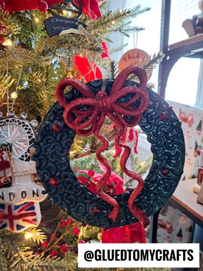 A dark green pasta noodle Christmas wreath ornament with red accents and a glittery red bow hangs on a Christmas tree, surrounded by other festive ornaments. The text “@GLUEDTOMYCRAFTS” appears at the bottom.