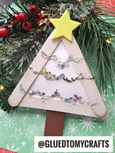 A handmade popsicle stick winter tree craft, decorated with glitter and star sequins, topped with a yellow star and set on a festive green and red background. The handle @GLUEDTOMYCRAFTS is visible.