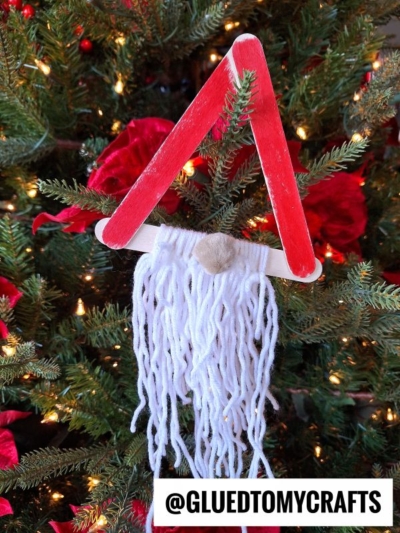 A yarn bearded Santa ornament made from red craft sticks in a triangle, with white yarn for the beard and a small nose at the top, hangs on a lit and decorated Christmas tree.