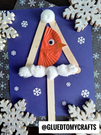 A winter-themed mixed media snowy birdhouse craft featuring a red cardinal made from a cupcake liner, with a googly eye and orange beak, perched on A-frame craft sticks, decorated with cotton balls and snowflakes on dark paper.