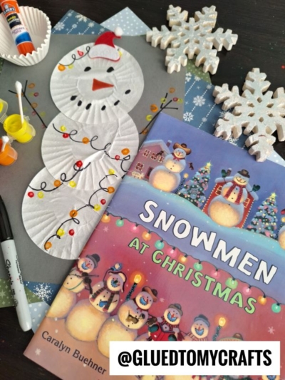 Christmas craft scene featuring adorable cupcake liner snowmen, the Snowmen at Christmas book, paint, glue, markers, snowflake decorations, and festive cupcake liners arranged on a dark surface. @GLUEDTOMYCRAFTS label at the bottom.