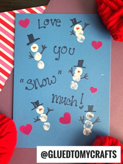 A blue fingerprint snowman card featuring white snowmen with black hats and orange noses, handwritten "Love you snow much!", and pink hearts. Red knitted fabric peeks from the corners for a cozy touch.