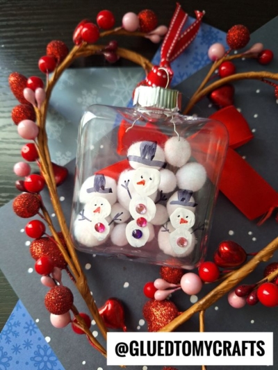 A clear ornament filled with white pom-poms, decorated as snowmen, sits on dark paper with a heart-shaped wreath of red and pink berries—perfect alongside fingerprint snowman cards. A red ribbon and the handle @GLUEDTOMYCRAFTS are visible.