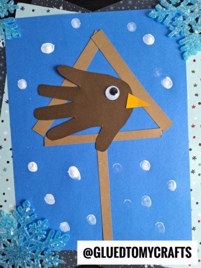 A mixed media snowy birdhouse craft for kids featuring a brown handprint bird with a yellow beak and googly eye, perched on a stick inside a triangle, set against a blue snowy background with paper snowflakes.