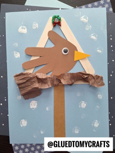 A kids craft features a brown handprint cutout shaped like a bird on a mixed media snowy birdhouse, with a yellow beak, googly eye, and crumpled brown paper perch, all set against a blue snowy background.