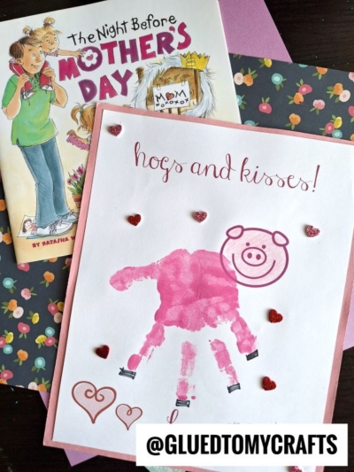 A handmade Mother's Day card with a pink handprint pig, heart stickers, and “hogs and kisses!” sits next to the children’s book The Night Before Mother’s Day. The background features floral and purple papers.