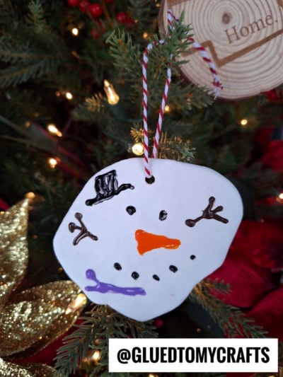 A handmade clay melting snowman face ornament with a painted hat, eyes, mouth, and carrot nose hangs on a Christmas tree. A wood slice decor piece and gold decorations are in the background. Text reads @GLUEDTOMYCRAFTS.