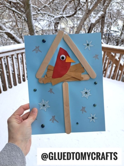A hand holds a mixed media snowy birdhouse craft featuring a red bird made of colored paper and a googly eye, perched in a craft stick house with snowflake stickers. Snowy trees and a wooden railing are visible in the background.