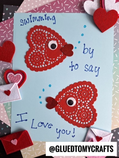 Two red heart-shaped doilies with googly eyes and fins are crafted as paper doily fish valentines on blue paper, surrounded by felt hearts and envelopes. Handwritten text reads “swimming by to say I love you!”.